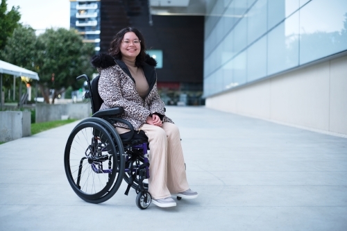 Smiling woman with a disability sitting in a wheelchair outside in a city - Australian Stock Image