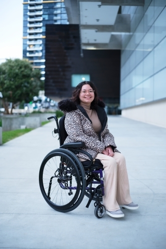 Smiling woman with a disability sitting in a wheelchair outside in a city - Australian Stock Image