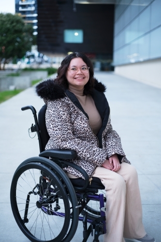 Smiling woman with a disability sitting in a wheelchair outside in a city - Australian Stock Image
