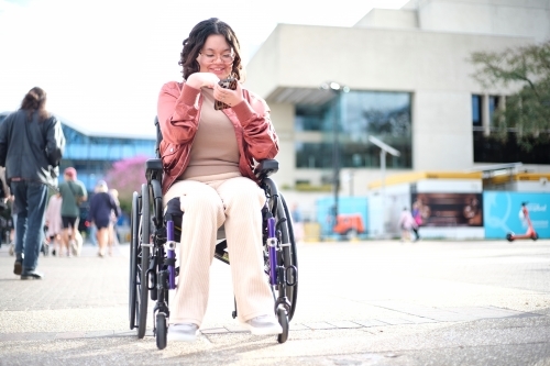 Smiling woman with a disability sitting in a wheelchair outside the city with mobile phone - Australian Stock Image