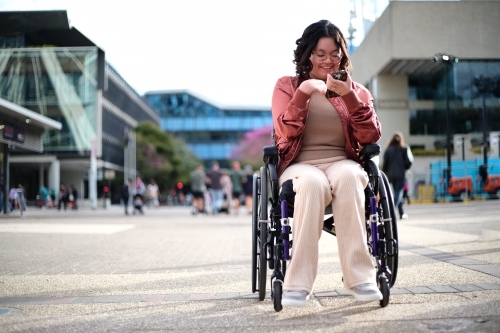 Smiling woman with a disability sitting in a wheelchair outside the city with mobile phone