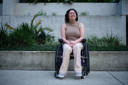 Smiling woman with a disability sitting in a wheelchair outside with grass and plants behind her - Australian Stock Image