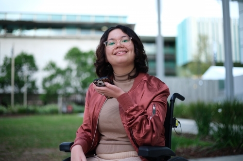 Smiling woman with a disability wearing glasses sitting in a wheel chair holding a mobile phone - Australian Stock Image