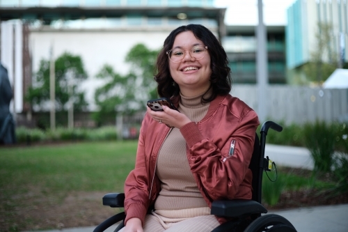 Smiling woman with a disability wearing glasses sitting in a wheel chair holding a mobile phone