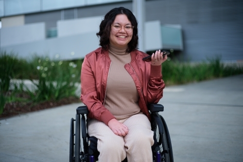 Smiling woman with a disability wearing glasses sitting in a wheel chair holding a mobile phone - Australian Stock Image