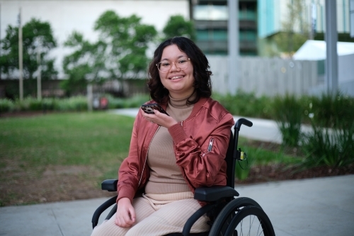 Smiling woman with a disability wearing glasses sitting in a wheel chair holding a mobile phone - Australian Stock Image
