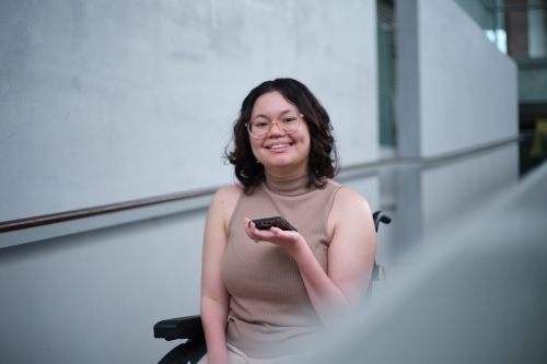 Smiling woman with a disability wearing glasses sitting in a wheel chair holding a mobile phone
