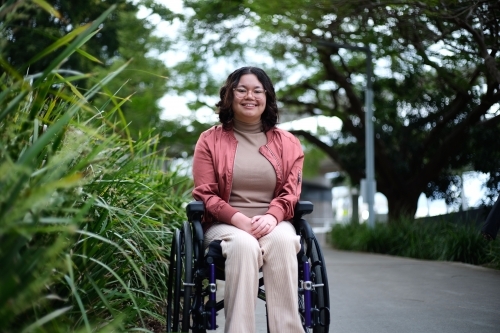 smiling woman with disability sitting in a wheelchair outside next to a tall grass - Australian Stock Image
