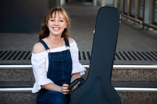 Smiling young adult with guitar case sitting on steps - Australian Stock Image