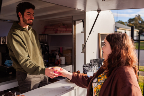smiling young Aussie barista man handing customer cup from coffee van in morning sunlight - Australian Stock Image