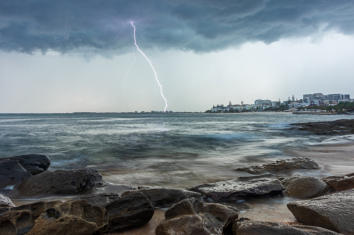 Storm clouds and lightning strike over ocean with rocky shoreline and coastal town in background - Australian Stock Image
