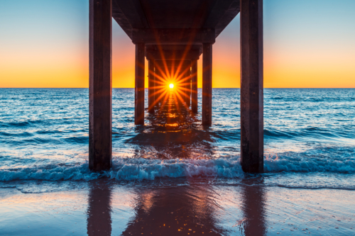 Sun shining through Brighton Jetty pylons while viewed from the beach at sunset time - Australian Stock Image