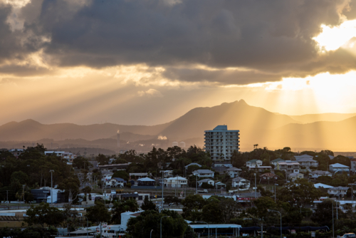 Sunbeams breaking through clouds over mountain silhouette and coastal town buildings at sunset - Australian Stock Image