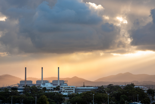 Sunlight breaking through storm clouds over city landscape and industrial smokestacks at sunset - Australian Stock Image
