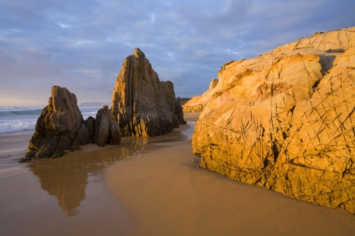 Sunlit yellow coastal rocks at sunrise with sand and cloud pattern