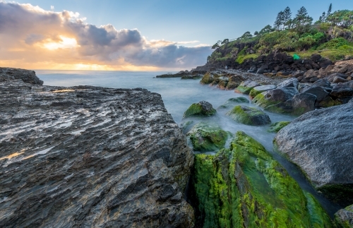 Sunrise at beach with waves crashing over green mossy rocks