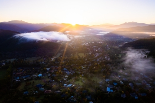Sunrise beams through the fog looking down onto a village filled with autumn trees - Australian Stock Image