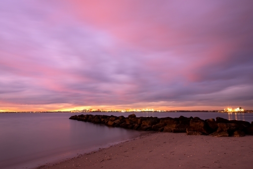 Sunset with lights from Port Botany in the distance and a beach with rock groyne in the foreground