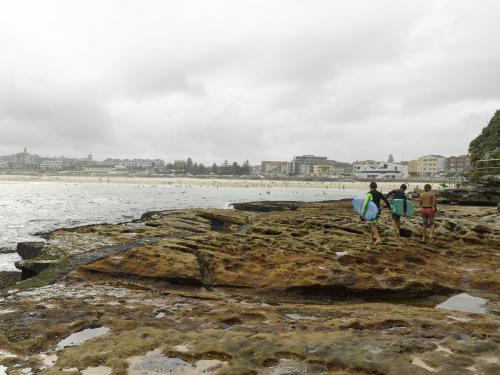 Surfer boys walking on rocks at Bondi Beach on an overcast day - Australian Stock Image