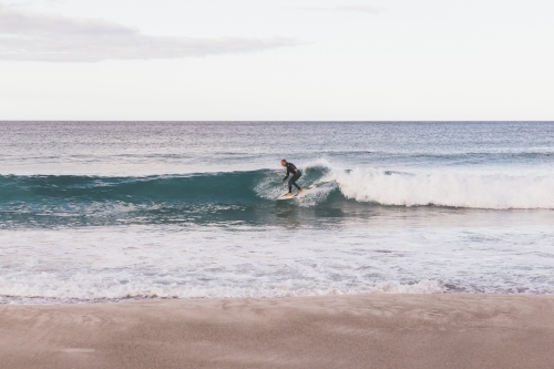 Surfer catching a wave in the ocean, light blue sky and grey cloud and sand in the foreground - Australian Stock Image