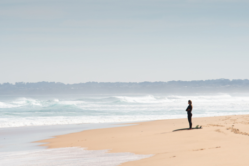 Surfer with board standing on beach looking out to the ocean - Australian Stock Image