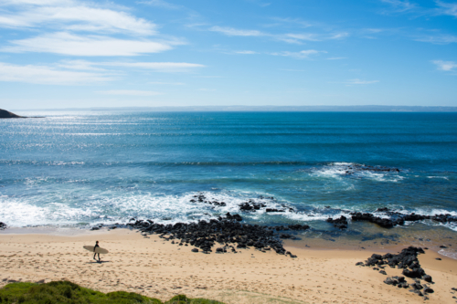 Surfer with board walking on beach with blue ocean in background - Australian Stock Image