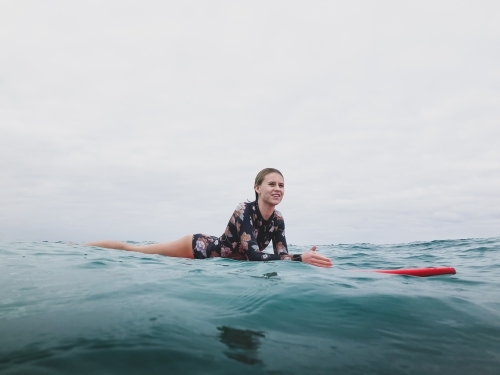 Surfing Teenage girl lying on red surfboard in ocean smiling wearing floral swimsuit on overcast day - Australian Stock Image
