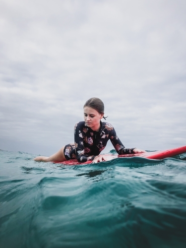 Surfing Teenage girl lying on red surfboard in ocean wearing floral swimsuit on overcast day
