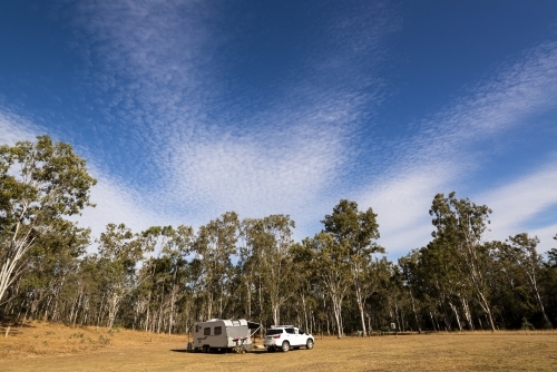SUV and caravan camped in a forest clearing under blue sky with radiating cloud patterns - Australian Stock Image