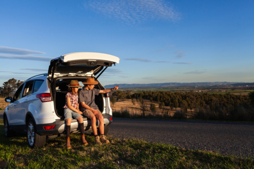 Teen boy and preteen girl siblings sitting in car boot beside rural Australian road in warm sunlight - Australian Stock Image