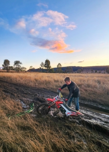 Teen boy on farm with motorbike stuck in the mud - Australian Stock Image