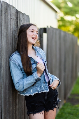 Teen girl laughing leaning on wooden fence in afternoon - Australian Stock Image