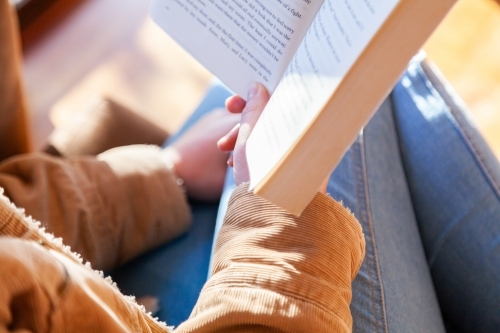 Teen girl reading a novel in the lounge room by a window in winter - Australian Stock Image