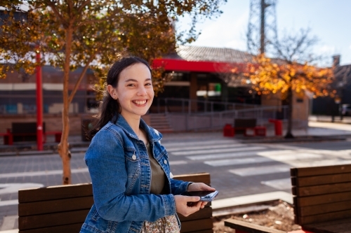 Teen girl with big smile holding device while outside on main street of country town - Australian Stock Image