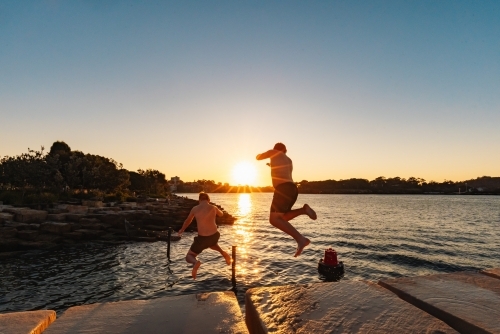 Teenage boy jumping from ledge into water in bright setting sun - Australian Stock Image