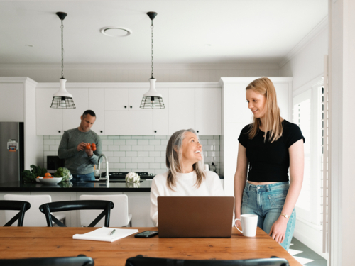 Teenage daughter giving mum a cup of coffee working on her laptop on wooden table - Australian Stock Image