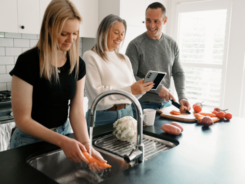 Teenage daughter helping dad prepare food by washing vegetables while mum on the phone - Australian Stock Image