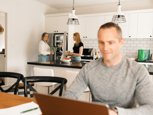 Teenage daughter helping mum with groceries in the kitchen while dad works on computer - Australian Stock Image