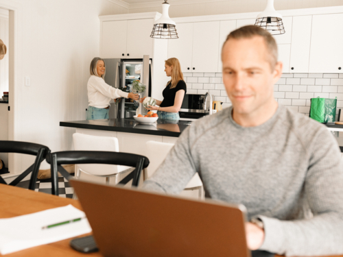 Teenage daughter helping mum with groceries in the kitchen while dad works on computer - Australian Stock Image