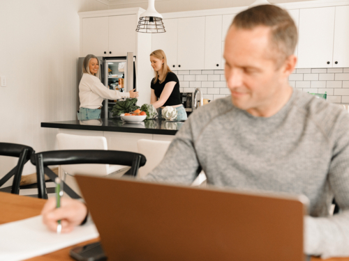 Teenage daughter helping mum with groceries in the kitchen while dad works on computer - Australian Stock Image