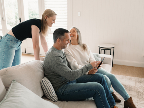 Teenage daughter leaning over her parents seated on the lounge looking at fold phone screen - Australian Stock Image