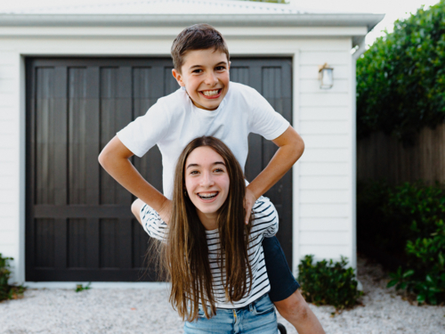 Teenage girl doing piggyback with brother on her back in their yard. - Australian Stock Image