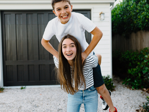 Teenage girl giving her brother a piggyback around their yard at home - Australian Stock Image