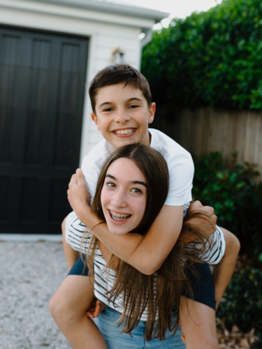 Teenage girl giving her brother a piggyback around their yard at home - Australian Stock Image
