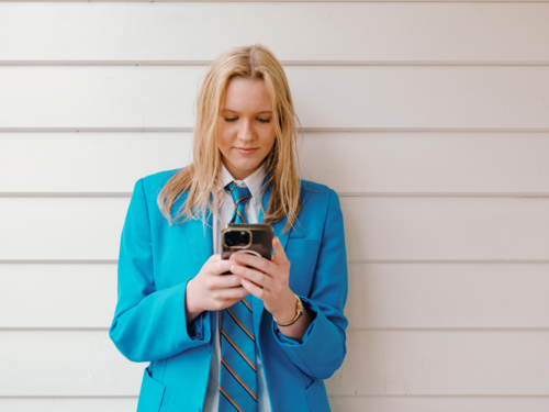 Teenage girl in school uniform leaning against the white wall while engaged on her phone - Australian Stock Image