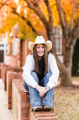 Teenage girl sitting on brick wall laughing wearing hat and scarf - Australian Stock Image