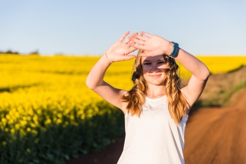 Teenage girl standing next to canola field crop shielding face with hands from the sun
