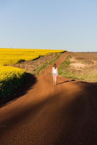 Teenage girl walking down dirt road on farm with canola crop - Australian Stock Image