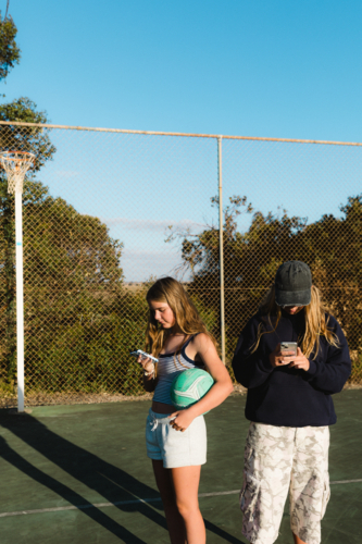 Teenage girls on smart phones while holding netball at outdoor court - Australian Stock Image
