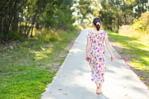 Teenage woman in floral dress in early Spring walking along a footpath through park in Singleton - Australian Stock Image